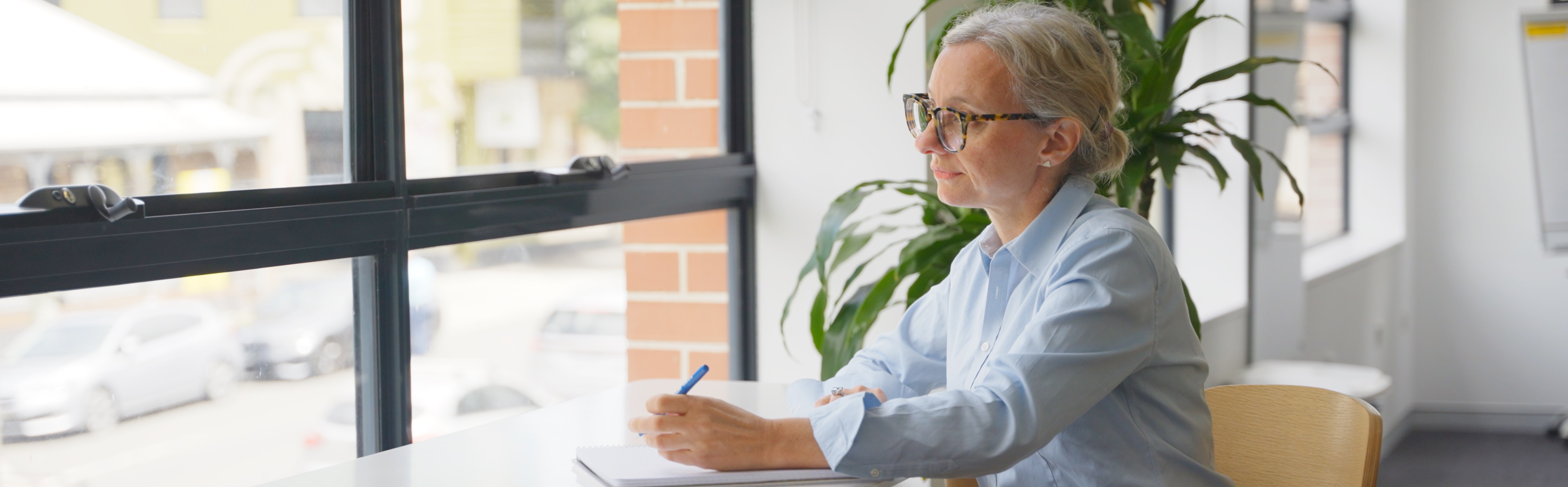 Woman with grey hair and glasses writing in a notebook at a bright, modern office desk near a window.